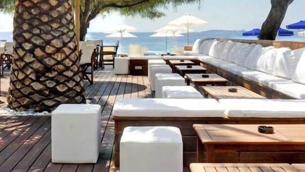 Une terrasse en bois avec des sièges et des tables rembourrés de couleur blanche, donnant sur une plage parsemée de parasols et de palmiers.
