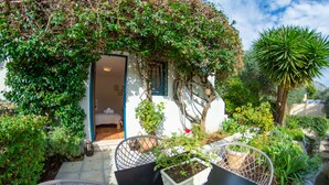 Un petit bâtiment blanc, doté d’une porte et d’une fenêtre bleues, entouré d’une végétation luxuriante et de plantes en pot.