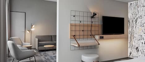 Una camera d'albergo moderna con un divano grigio, un piccolo tavolino rotondo, una poltrona grigia, una TV montata su una parete a griglia e carta da parati decorata.