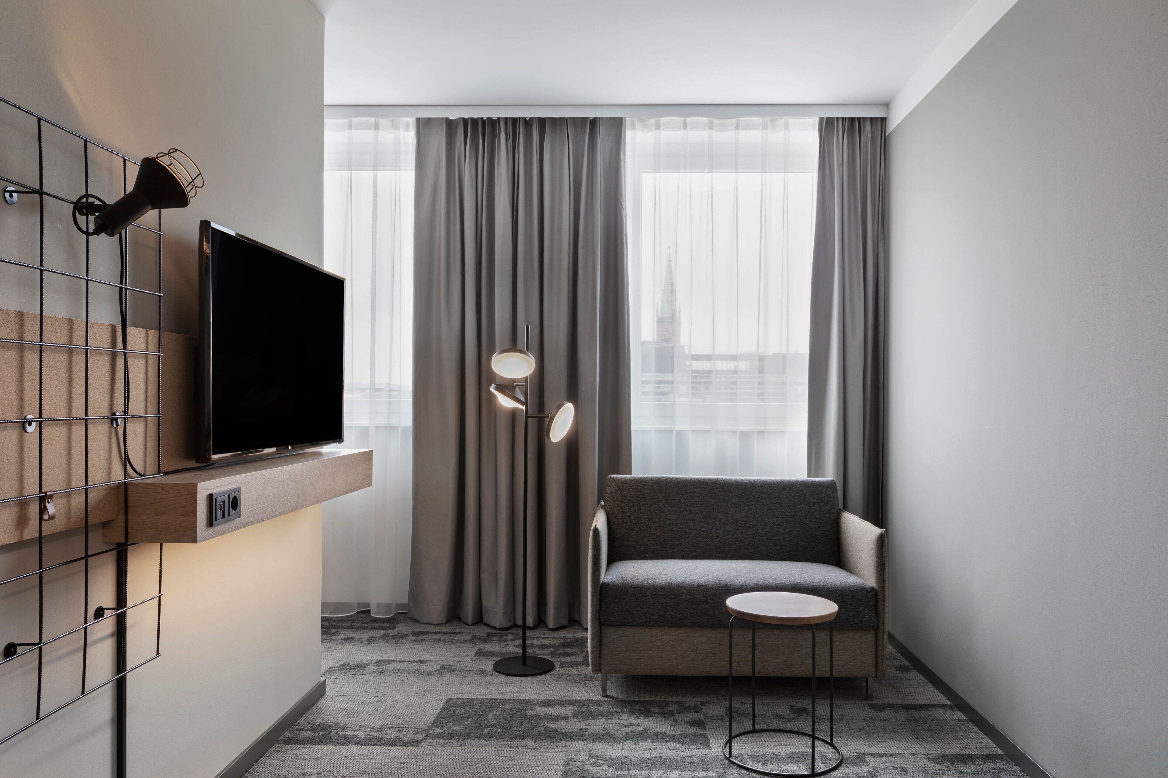 A modern hotel room with a flat-screen TV mounted on a wooden panel, a grey sofa, a small round table, and a floor lamp.