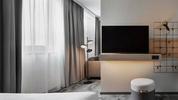 A modern hotel room with a large bed, a TV mounted on the wall, a small round ottoman, and a floor lamp.