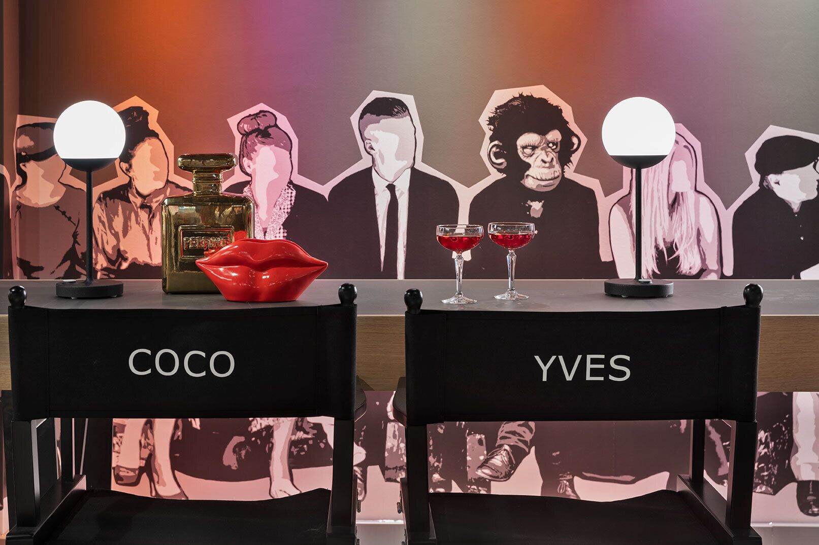 Una zona de bar con dos sillas con los nombres 'COCO' e 'YVES', un cuenco con forma de barra de labios rojo, dos vasos de bebida roja y una botella de licor.