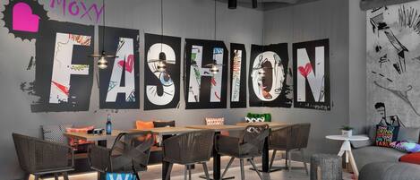 A modern interior with a large wall mural spelling "MOXY FASHION," a wooden table with chairs, and a colorful rug.