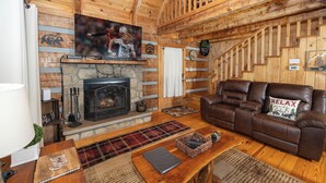 A cozy log cabin interior with a fireplace, leather sofa, and a television mounted above it. - Cozy Cabin - Large Yard w/ Firepit, walk to Fishing Pond, Pool, & Mini Golf (Sevierville)