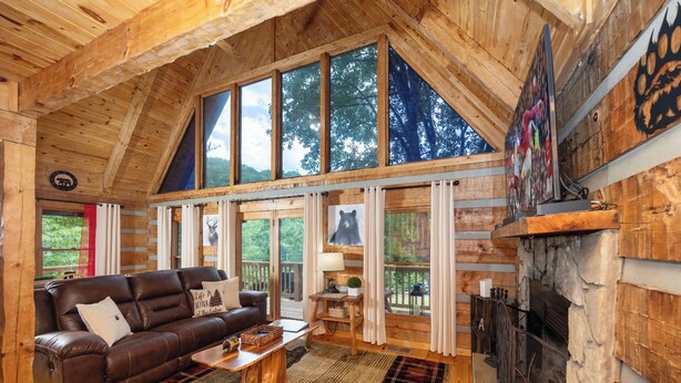 A log cabin interior with a leather sofa, a fireplace, and large windows. - Cozy Cabin - Large Yard w/ Firepit, walk to Fishing Pond, Pool, & Mini Golf (Sevierville)