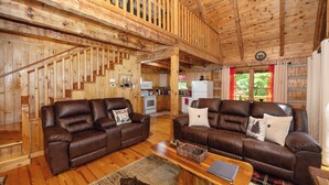 A wooden interior with two leather sofas, a wooden coffee table, and a kitchen area with a refrigerator and stove. - Cozy Cabin - Large Yard w/ Firepit, walk to Fishing Pond, Pool, & Mini Golf (Sevierville)