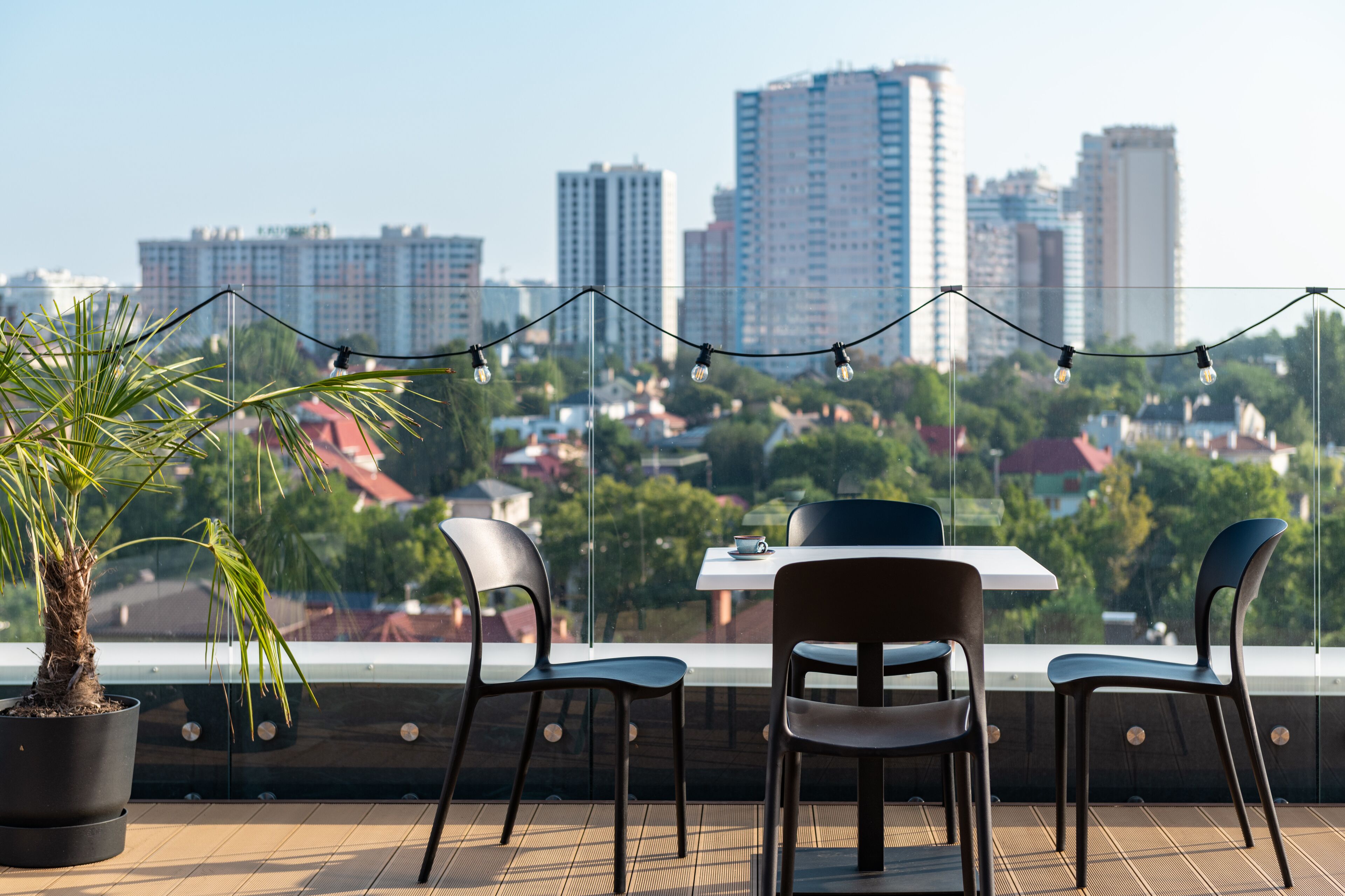Un balcón con mesa y sillas, una planta en maceta y vistas de un paisaje urbano con edificios altos.