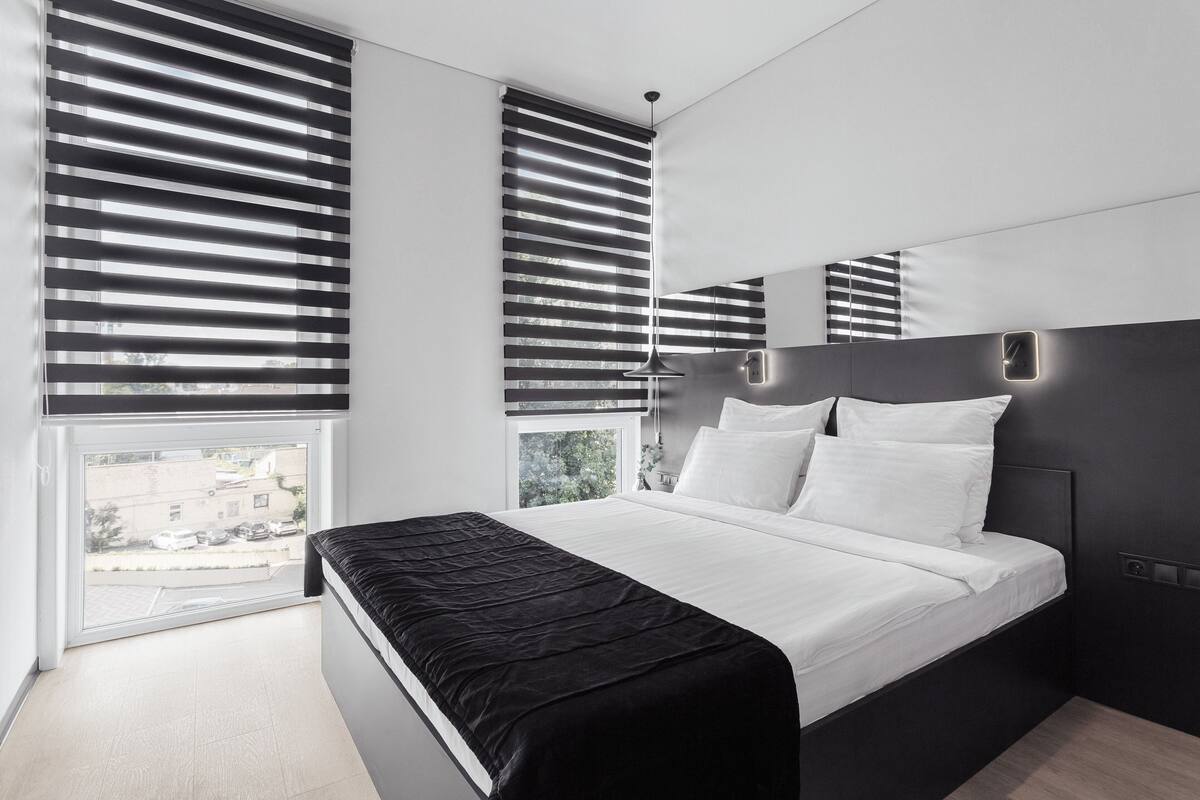 A modern bedroom with a large bed, black and white blinds, a black headboard, and a view of the outside.