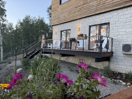 A modern house with a stone facade, a wooden deck with outdoor seating, and a small staircase leading to the deck.