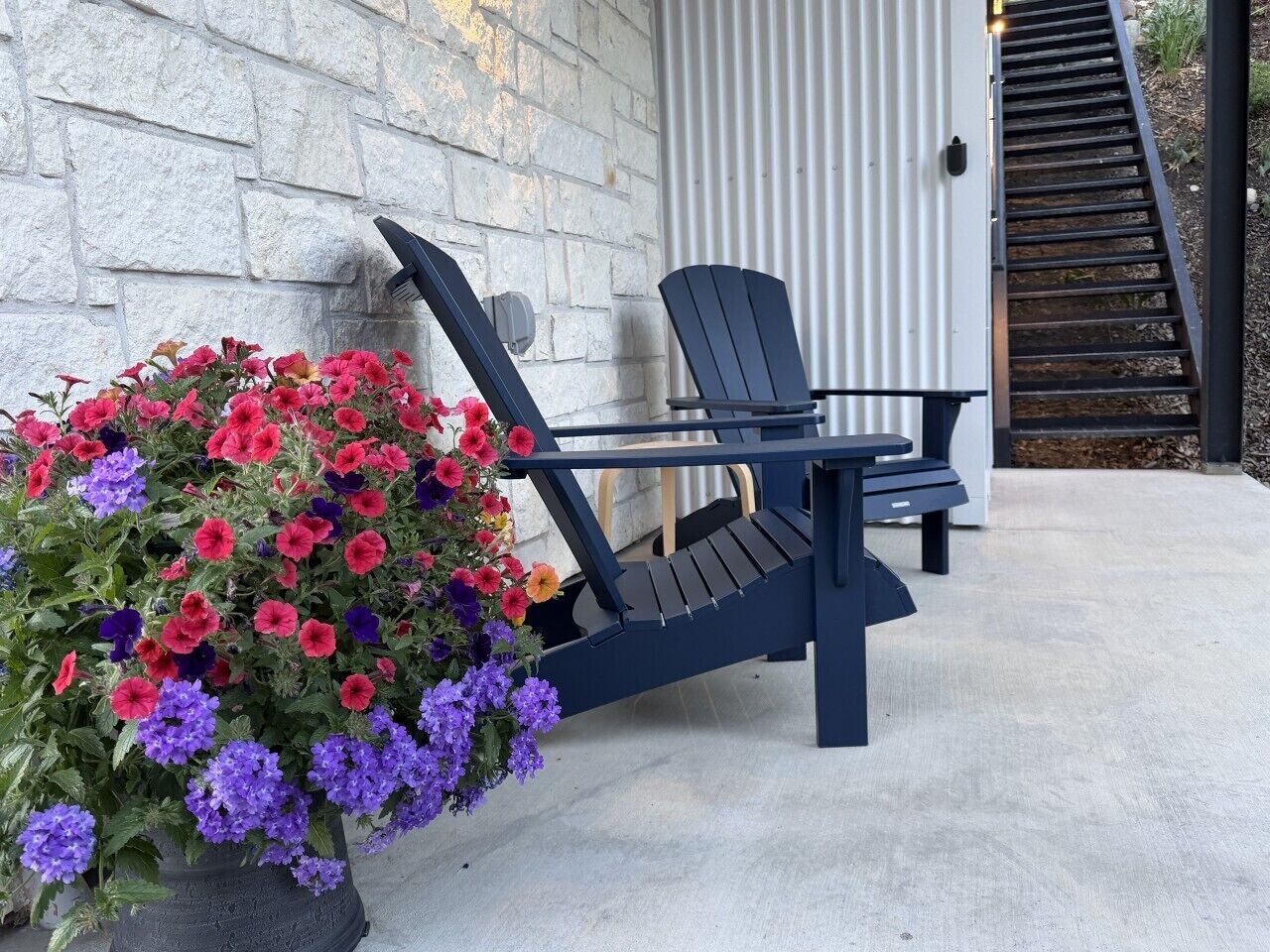 Um pátio de concreto com uma cadeira Adirondack preta e uma mesa, uma planta em vaso com flores vermelhas e roxas, e uma escadaria de metal.