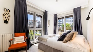 Une chambre moderne avec un grand lit, un fauteuil et une vue sur la ville grâce à de grandes fenêtres.