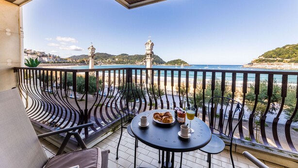 Un balcon avec une table dressée pour deux personnes, offrant une vue sur la côte, avec des bâtiments et une statue.