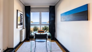 Une chambre moderne avec une table en verre, une chaise noire, un tableau encadré et une vue sur la mer grâce à de grandes fenêtres.
