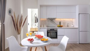 Ett modernt kök med vita köksskåp, en inbyggd ugn och ett kylskåp. Det finns ett matbord med stolar och en skål med frukt.