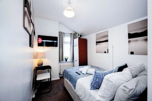 Ein Schlafzimmer mit Bett, Stuhl, Schreibtisch, Lampe, Heizkörper, einem Fenster mit Vorhängen und Bildern an den Wänden.