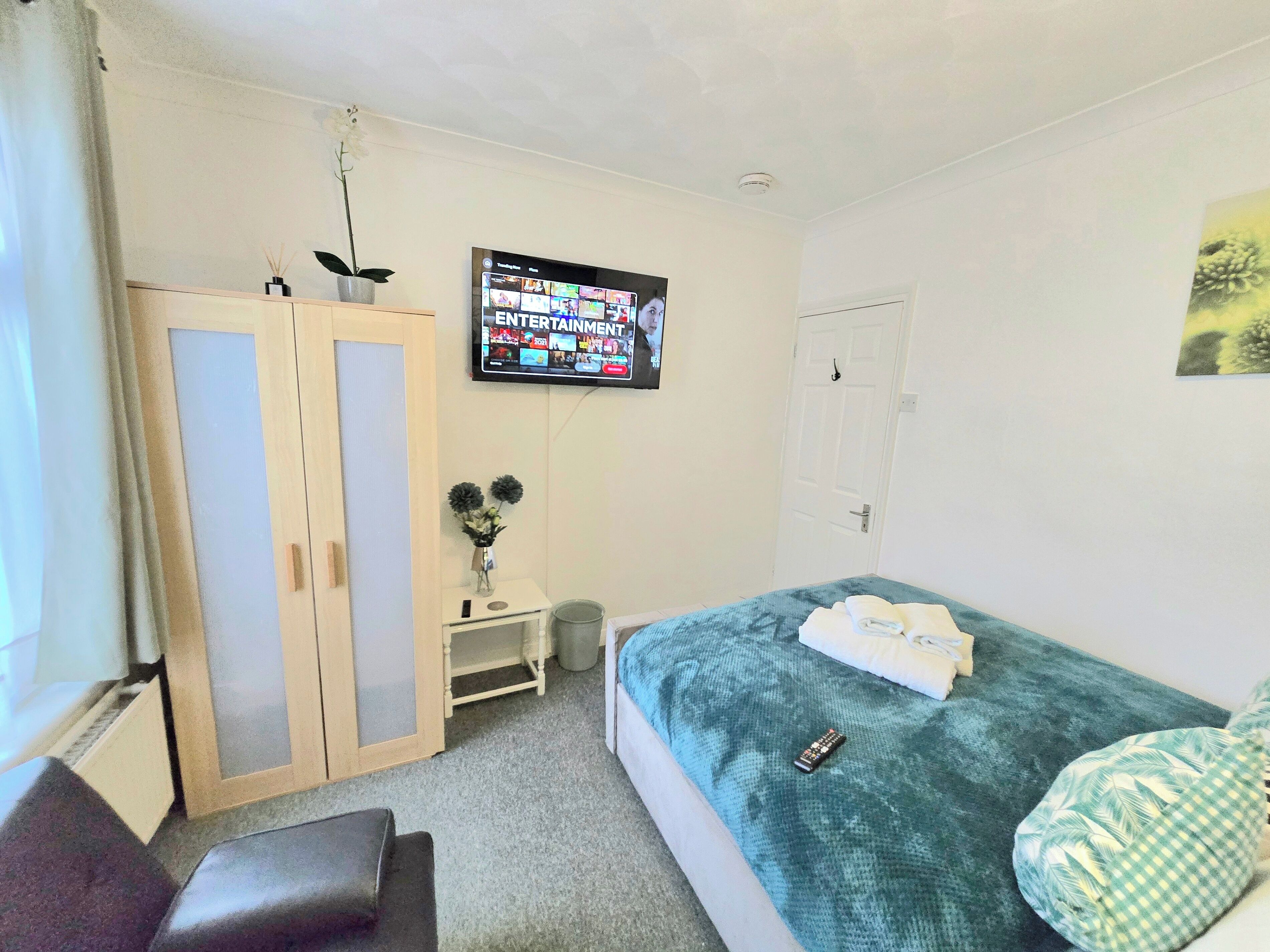 A hotel room with a bed, a TV mounted on the wall, a wardrobe, a small table with a vase, and a window with curtains.