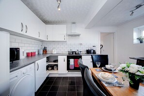 A modern kitchen with white cabinets, a dishwasher, a microwave, a stove, and a dining area with a table set for two. - Gilling Villa Remarkable 4-bed House in Gillingham (Gillingham)