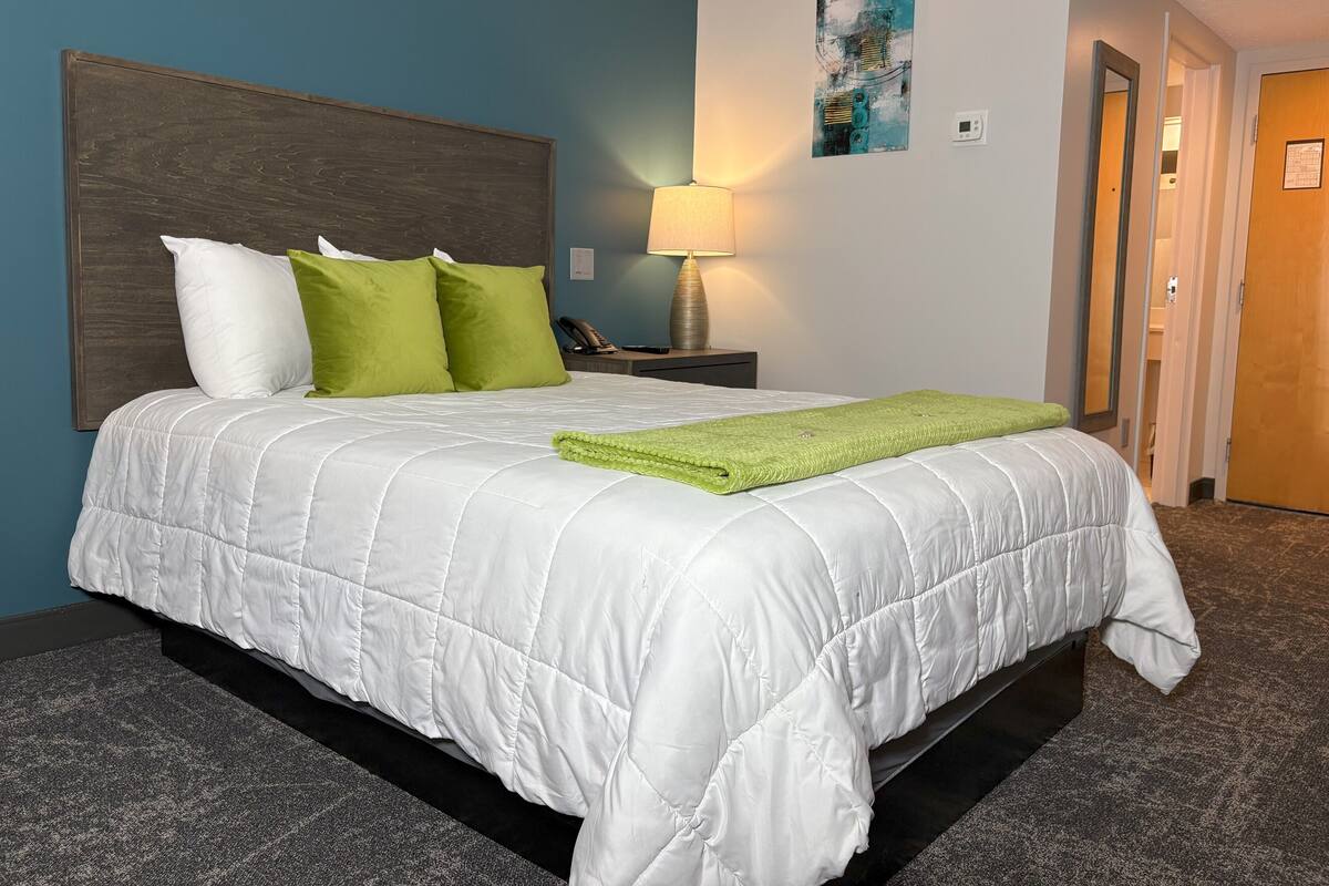 Una cama bien hecha con edredón blanco y manta verde, cabecera de madera, lámpara de noche y una obra de arte abstracta en la pared.