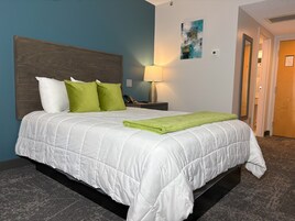 A neatly made bed with a white duvet and green throw, a wooden headboard, a bedside lamp, and a wall-mounted abstract art piece.