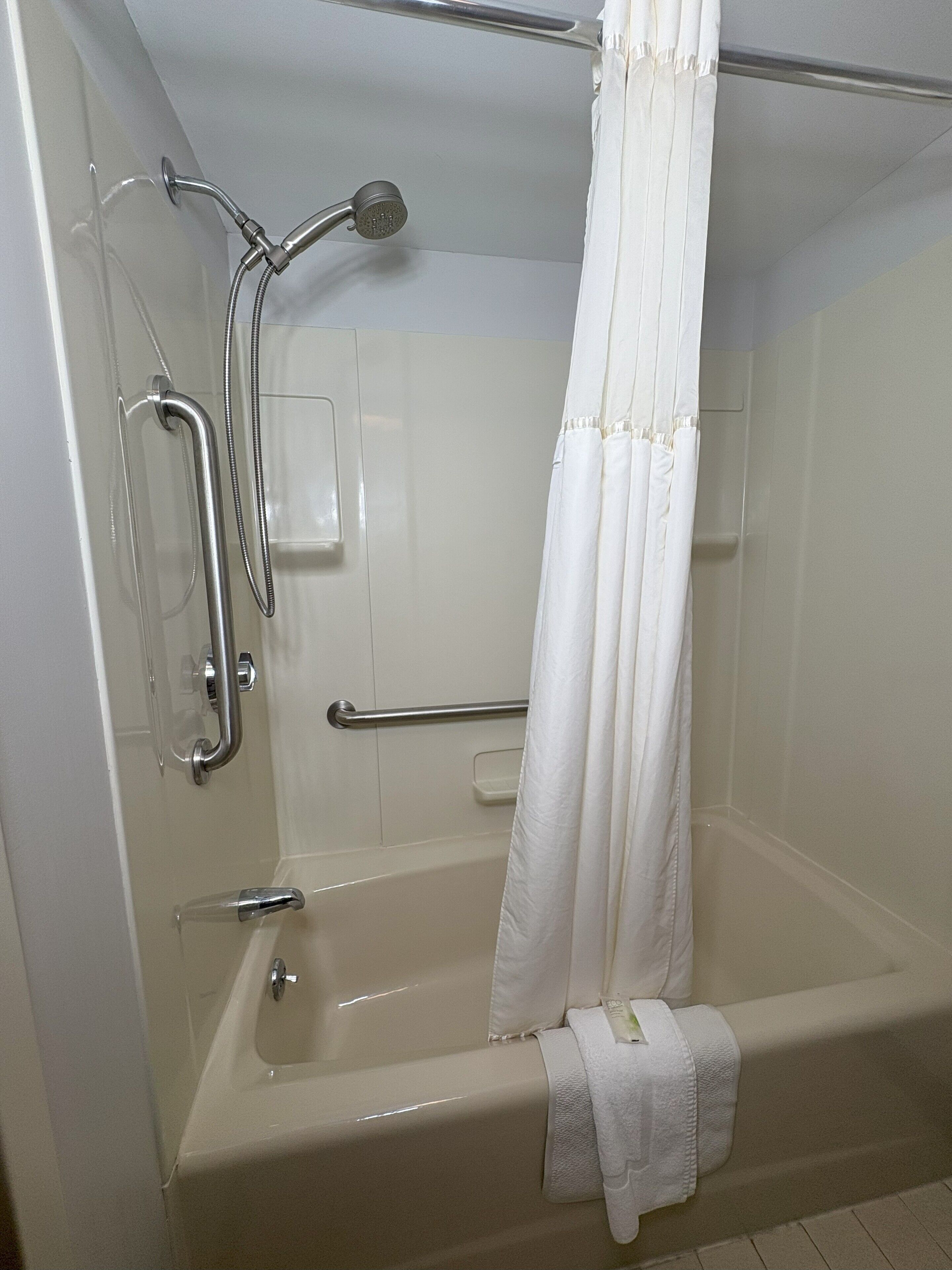 A modern bathroom with a walk-in shower, grab bars, and a white towel on the floor.