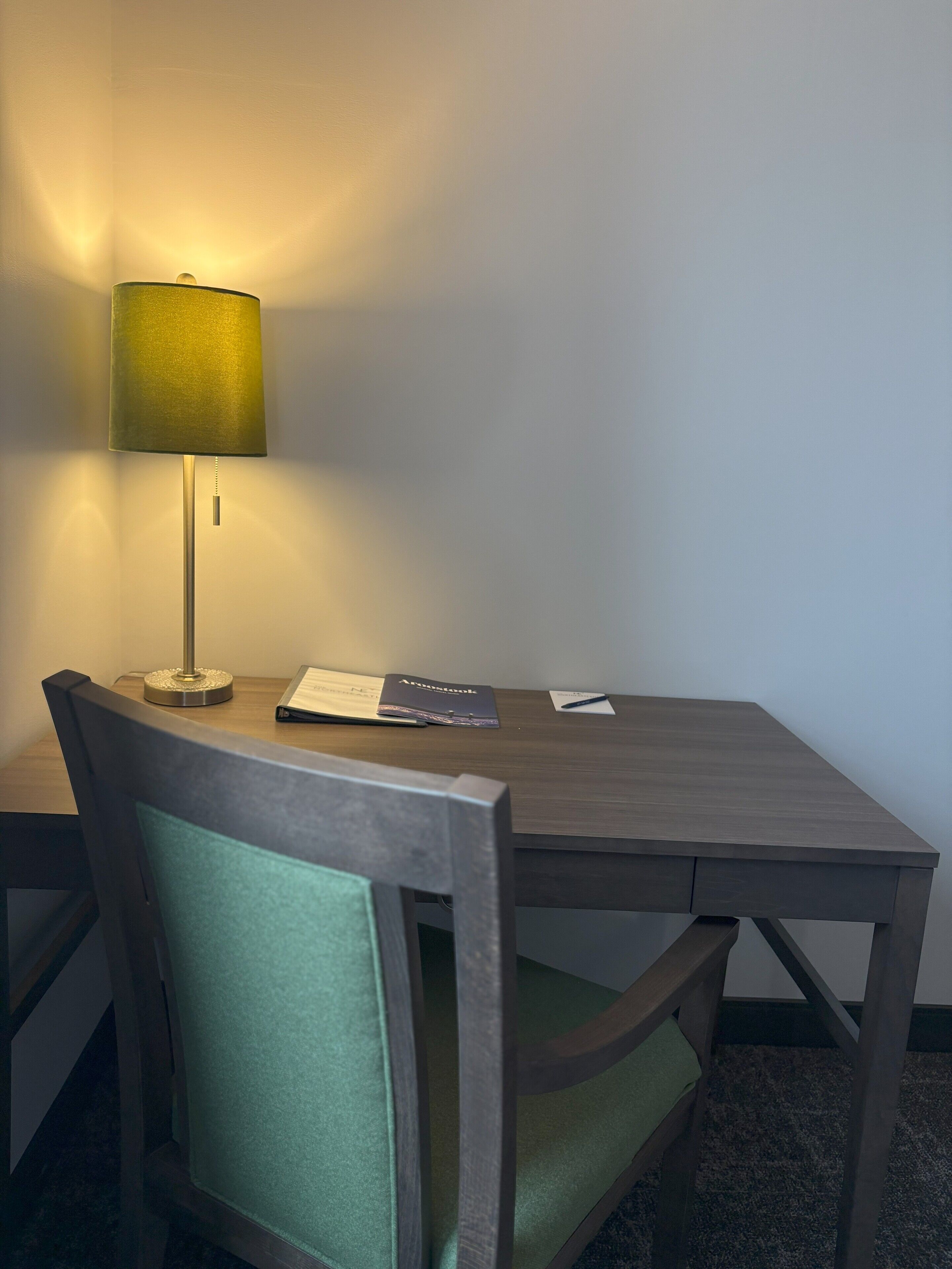 A hotel room with a wooden table, a green cushioned chair, a lamp, and a few items on the table including a notebook and a pen.