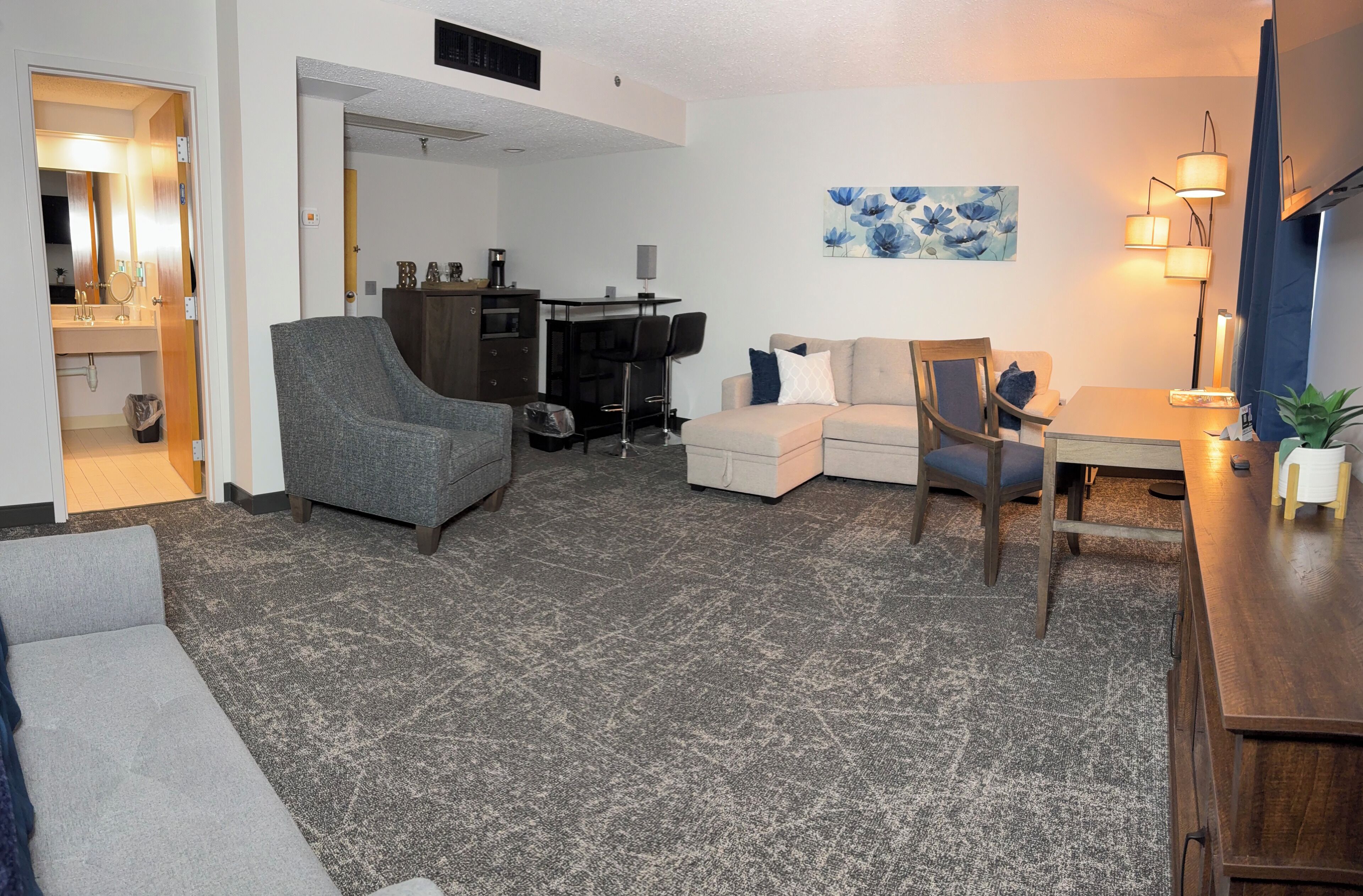 A living room with a grey armchair, a beige sofa, a wooden dining table, and a small kitchen area with a bar stool.