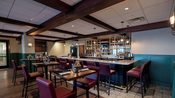 Zona de bar con mesas de madera y sillas rojas, una barra con diversas botellas y lámparas colgantes.