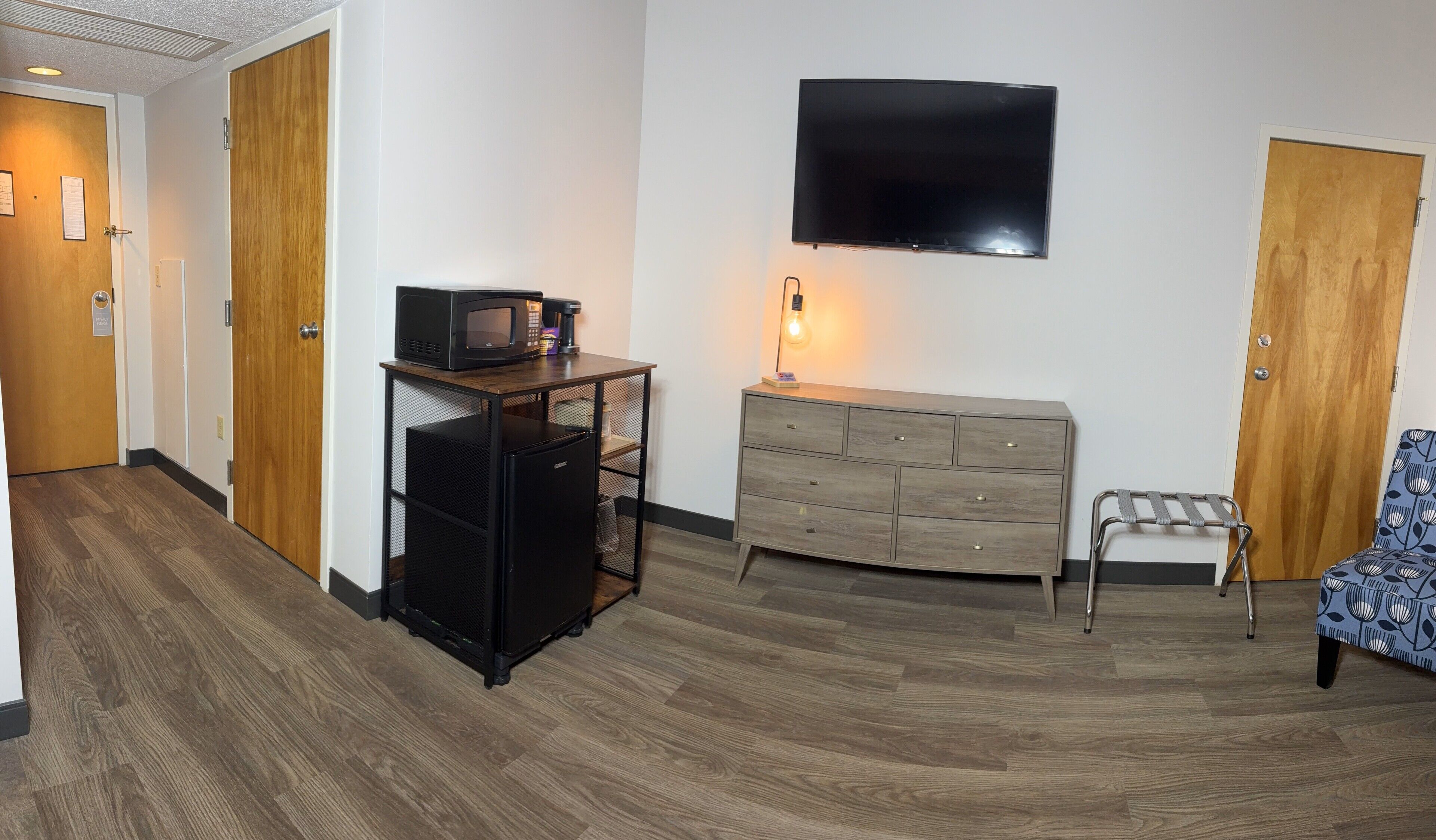 A hotel room with a wooden floor, a black refrigerator, a wooden cabinet, a TV mounted on the wall, and a chair with a floral pattern.