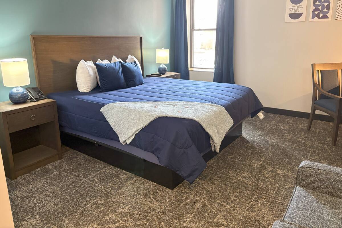 Habitación de hotel ordenada con una cama grande, una mesita de noche con lámpara, una silla y una obra de arte en la pared.