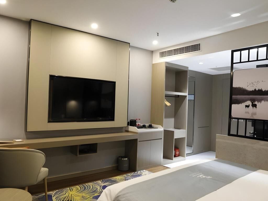 A modern hotel room with a large flat-screen TV mounted on the wall, a bed with a patterned bedspread, a small seating area with a chair, and a window with a view of a landscape.