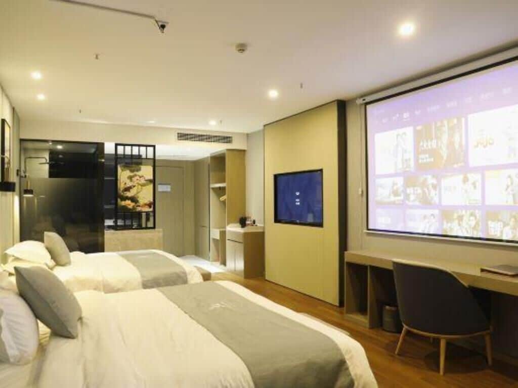 A hotel room with two beds, a desk, a TV, and a projection screen displaying images.