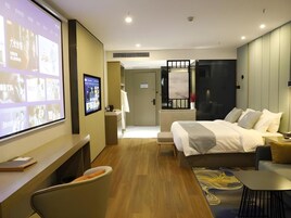 Una habitación de hotel moderna con cama, escritorio, silla y una pantalla de televisión mostrando imágenes.