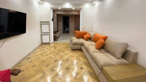 A living room with a flat-screen TV, a beige sofa with orange cushions, a wooden floor, and recessed ceiling lighting.