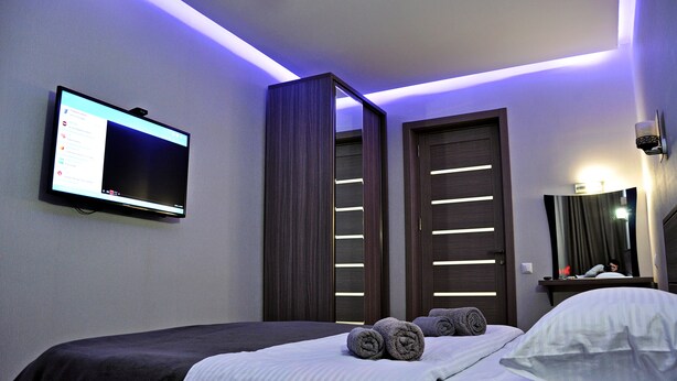 A modern hotel room with a bed, a flat-screen TV mounted on the wall, a wardrobe, and a mirror with a light.