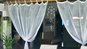 A room with a glass door, white curtains, a hanging dreamcatcher, a potted plant, and a wooden table with a bowl and a plate. - Pousada Arraial da Poesia (Porto Seguro)