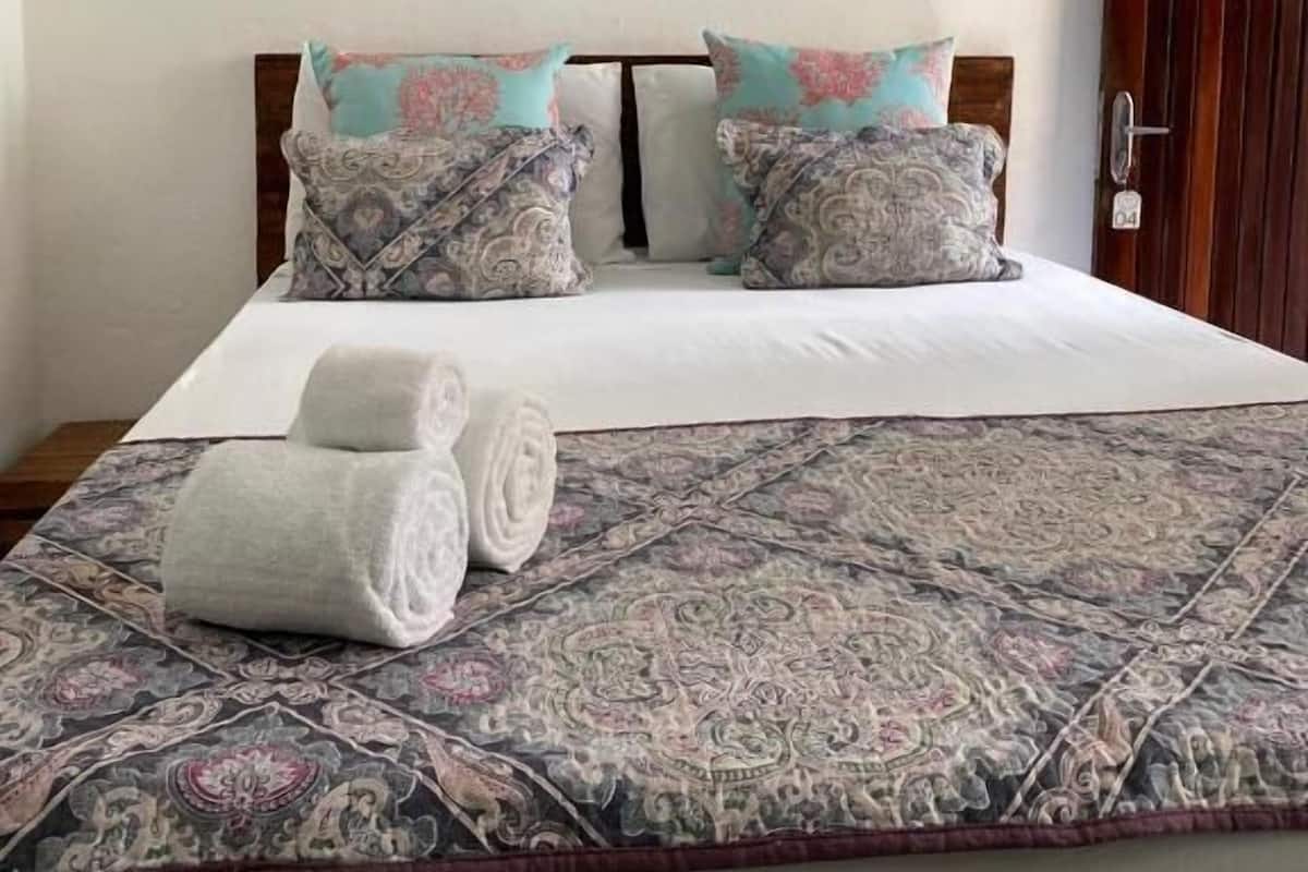 A neatly made bed with patterned bedding, two pillows, and rolled towels on a patterned rug.