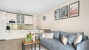 A modern living room with a grey sofa, a wooden coffee table, and a white rug. There is a kitchen area with white cabinets and a dining table with chairs. - Elite Apartments Mila (Gdansk)
