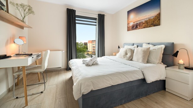 A modern hotel room with a bed, desk, chair, and a view of buildings through the window. - Elite Apartments Mila (Gdansk)