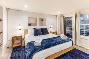 Une chambre à coucher bien rangée, avec un grand lit, deux tables de chevet avec des lampes, une fenêtre avec des rideaux et une vue sur un mur de briques à l’extérieur.