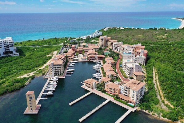 An aerial view of a coastal resort with multiple buildings, a marina with boats, and a clear view of the ocean.