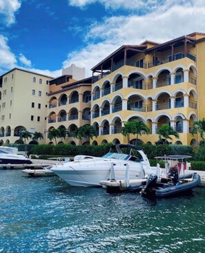 A marina with boats docked, a yellow building with balconies, and palm trees.