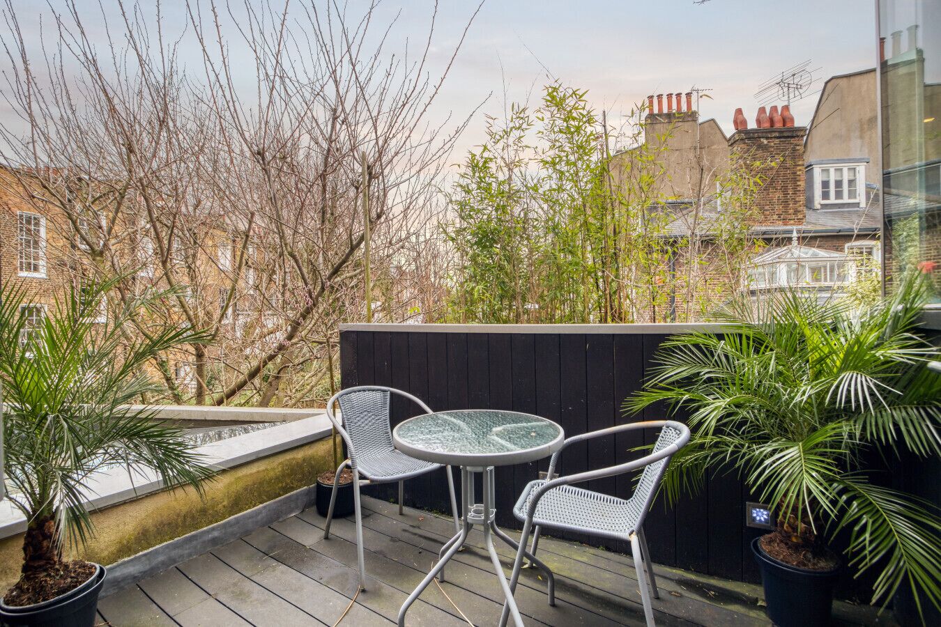 A balcony with a round glass table and two chairs, surrounded by potted plants and overlooking a cityscape.