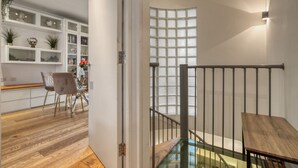 Ein modernes Interieur mit Holzboden, einer Essecke mit Stühlen, einer Treppe mit Glastrennwand und einer Wand mit dekorativen Gegenständen auf Regalen.
