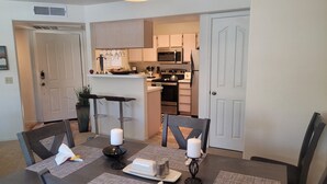 Un comedor con una mesa lista para cuatro, una cocina al fondo y una puerta que da a otra habitación.