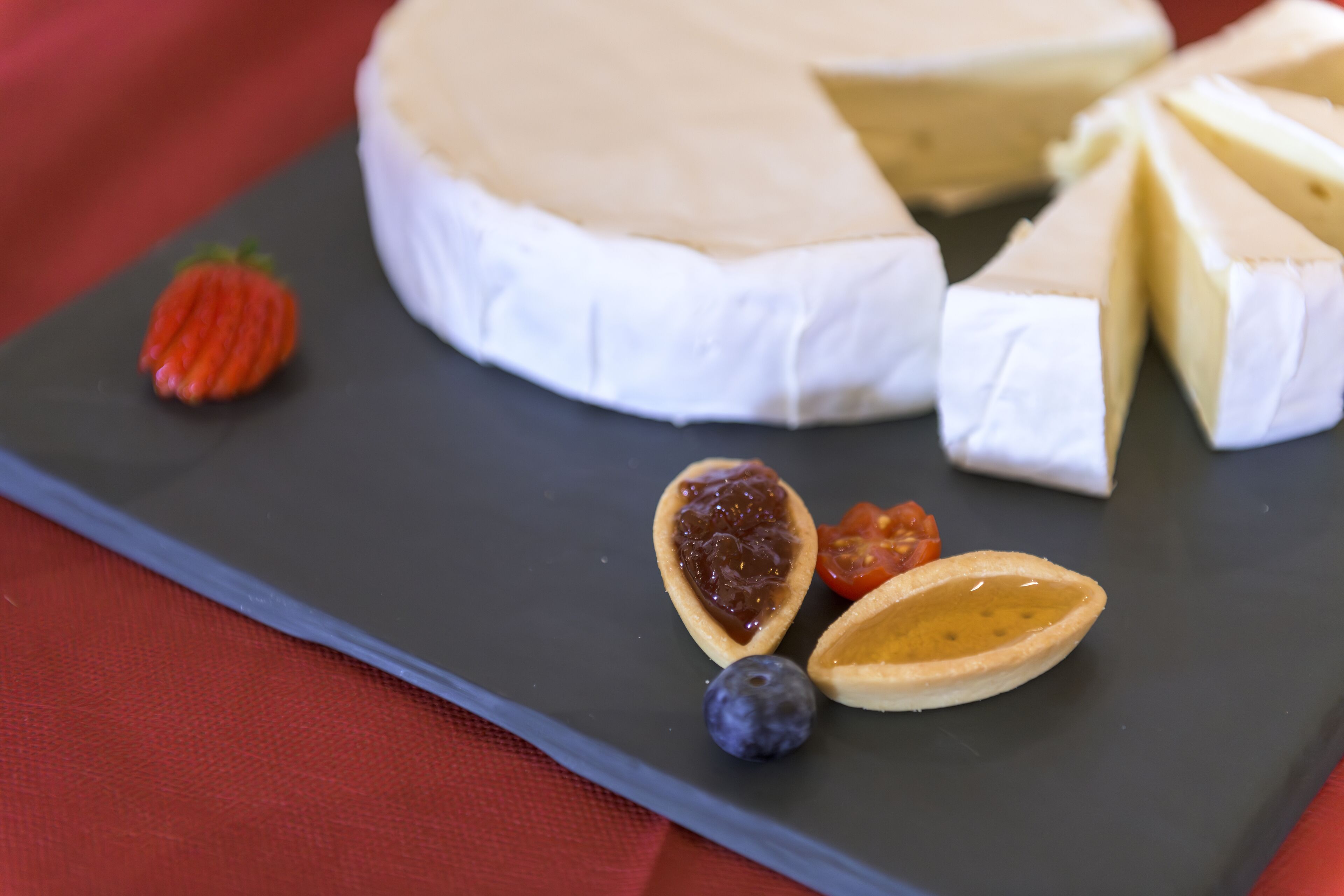 A round cheese wheel with a slice cut out, accompanied by a strawberry, a blueberry, and a fig.