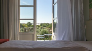 A hotel room with a bed, a window, and a view of a balcony with chairs and greenery.