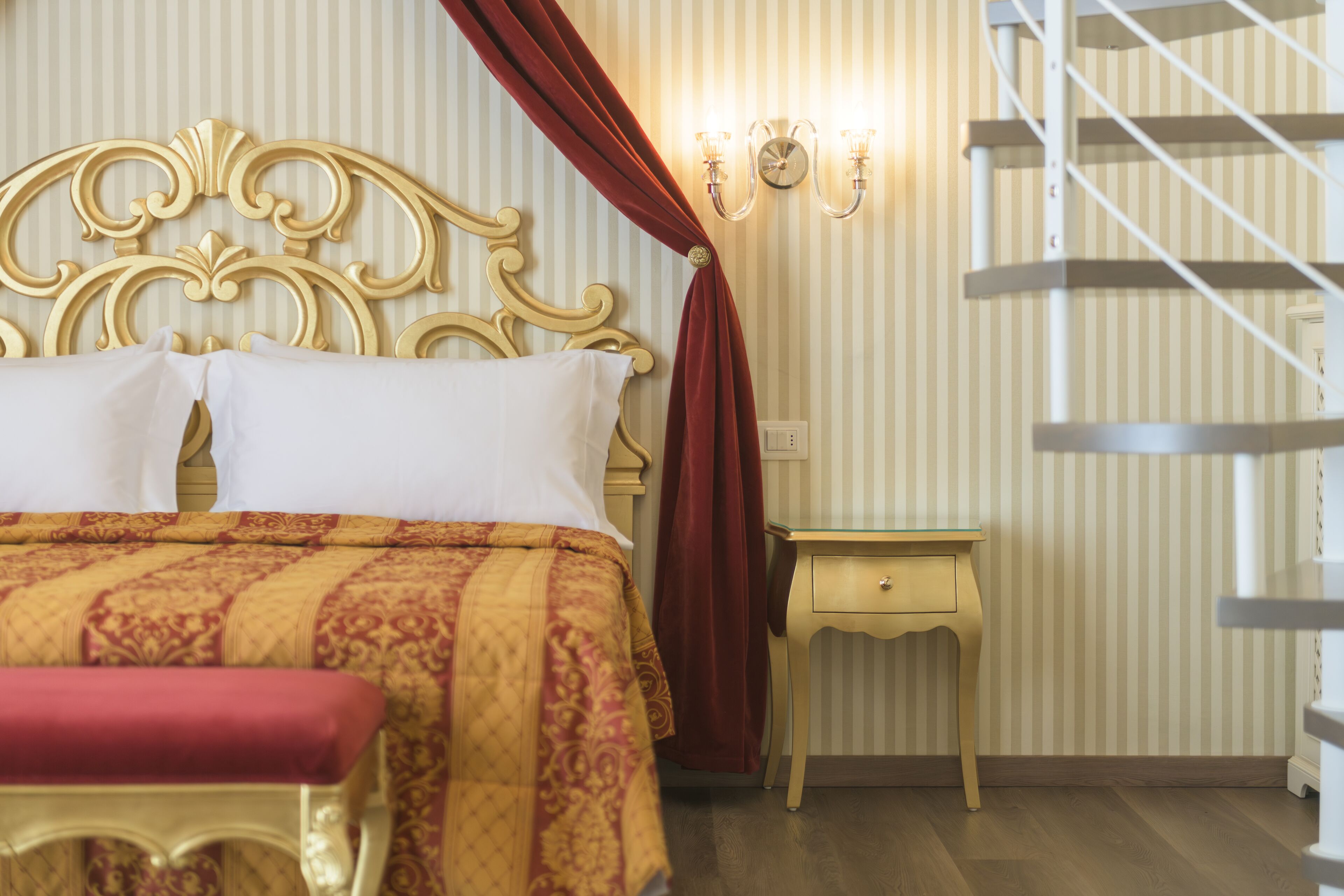 Una habitación de hotel con una cama grande, una mesita de noche, un banco rojo y una escalera.