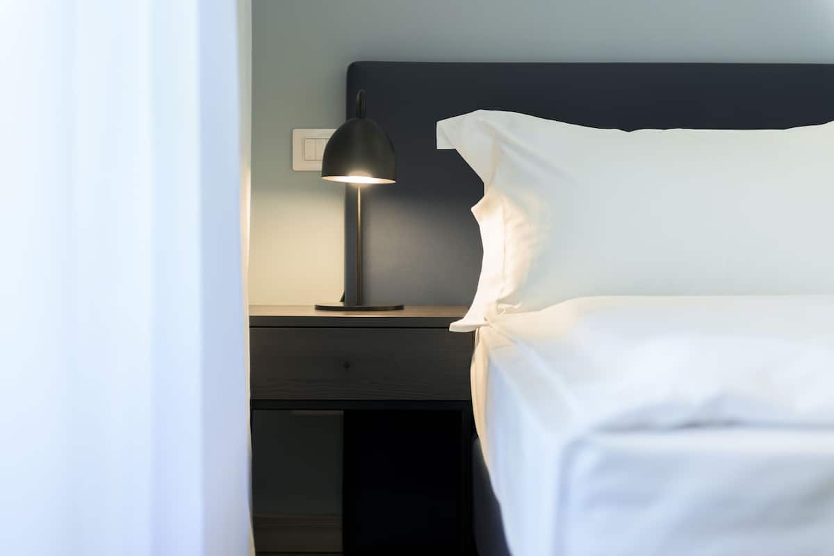 Una cama bien hecha con sábanas blancas, una mesita de noche negra con lámpara e interruptor de pared.