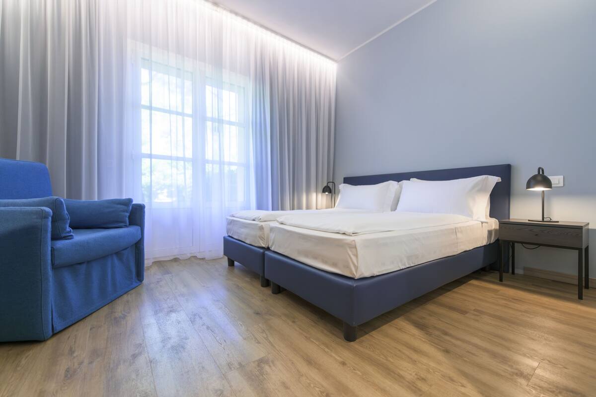 Una habitación de hotel moderna con una cama, un sofá azul, una mesita de noche y un ventanal con cortinas translúcidas.