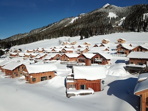 Ein verschneites Dorf mit Holzhäusern, Bergen im Hintergrund und blauem Himmel.